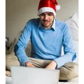 Man sitting on a bed at a laptop shopping with a Christmas hat on
